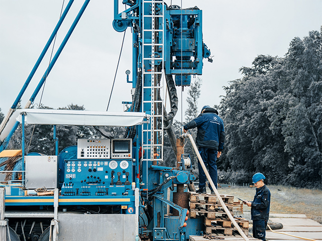 Auf dem Bild stehen zwei Männer in Schutzkleidung an einer großen, blau lackierten Bohranlage im Freien. Die Maschine ist komplex aufgebaut, mit vielen Hebeln, Anzeigen, Schläuchen und Metallteilen. Einer der Männer steht auf einem Stapel aus Holzpaletten und hält einen langen, hellen Schlauch, der in die Bohranlage geführt wird. Er trägt einen blauen Schutzhelm und eine dunkelblaue Arbeitsjacke. Der zweite Mann steht etwas abseits, ebenfalls mit Helm, und bedient offenbar ein mobiles Gerät oder schaut darauf. Im Hintergrund sind Bäume und ein grauer, bedeckter Himmel zu sehen, was der Szene eine kühle, industrielle Atmosphäre verleiht.