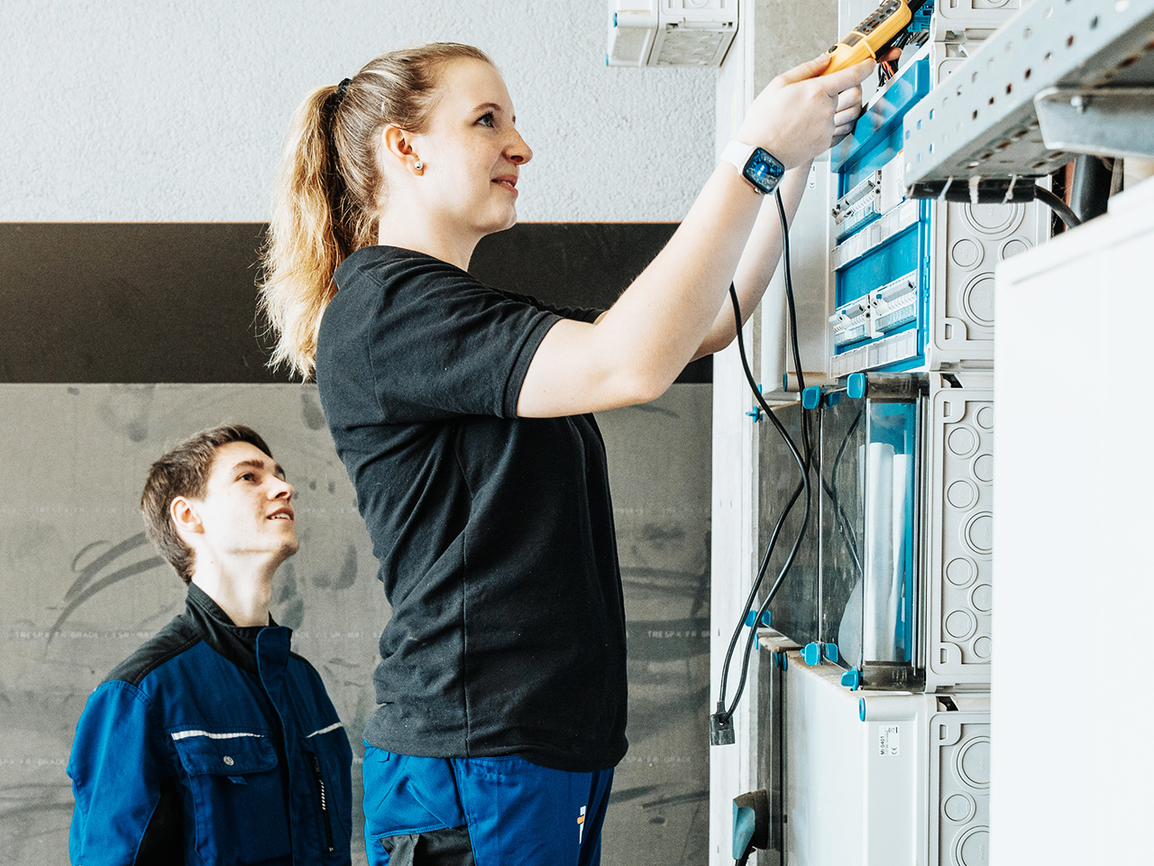 Im rechten Vordergrund steht eine Frau in schwarzem T-Shirt, die an einem hohen Schaltschrank arbeitet. Sie ist seitlich zu sehen, hebt beide Arme und hält in der rechten Hand ein Messgerät. Links hinter ihr steht ein junger Mann in blauer Arbeitskleidung, er schaut aufmerksam auf ihre Arbeit. Der Schaltschrank befindet sich rechts im Bild, mit mehreren Reihen von blauen und grauen Bauteilen. Der Hintergrund ist eine weiße Wand mit einem grauen horizontalen Streifen im oberen Bilddrittel.