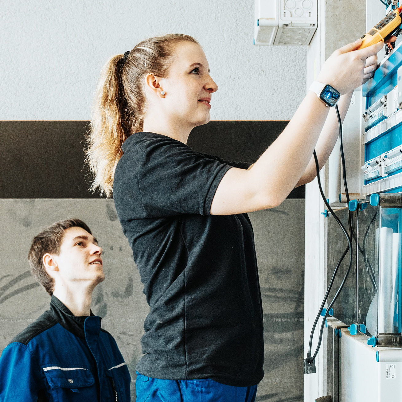 Im rechten Vordergrund steht eine Frau in schwarzem T-Shirt, die an einem hohen Schaltschrank arbeitet. Sie ist seitlich zu sehen, hebt beide Arme und hält in der rechten Hand ein Messgerät. Links hinter ihr steht ein junger Mann in blauer Arbeitskleidung, er schaut aufmerksam auf ihre Arbeit. Der Schaltschrank befindet sich rechts im Bild, mit mehreren Reihen von blauen und grauen Bauteilen. Der Hintergrund ist eine weiße Wand mit einem grauen horizontalen Streifen im oberen Bilddrittel.