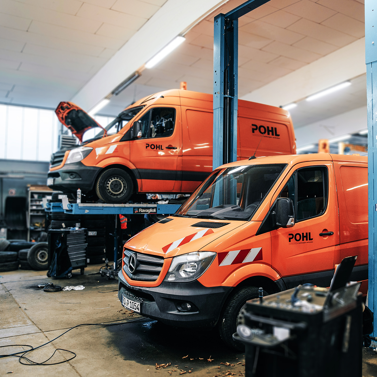 In einer Werkstatt stehen zwei orange POHL-Transporter auf Hebebühnen; Motorhauben geöffnet, daneben Werkzeugwagen und Reifen.