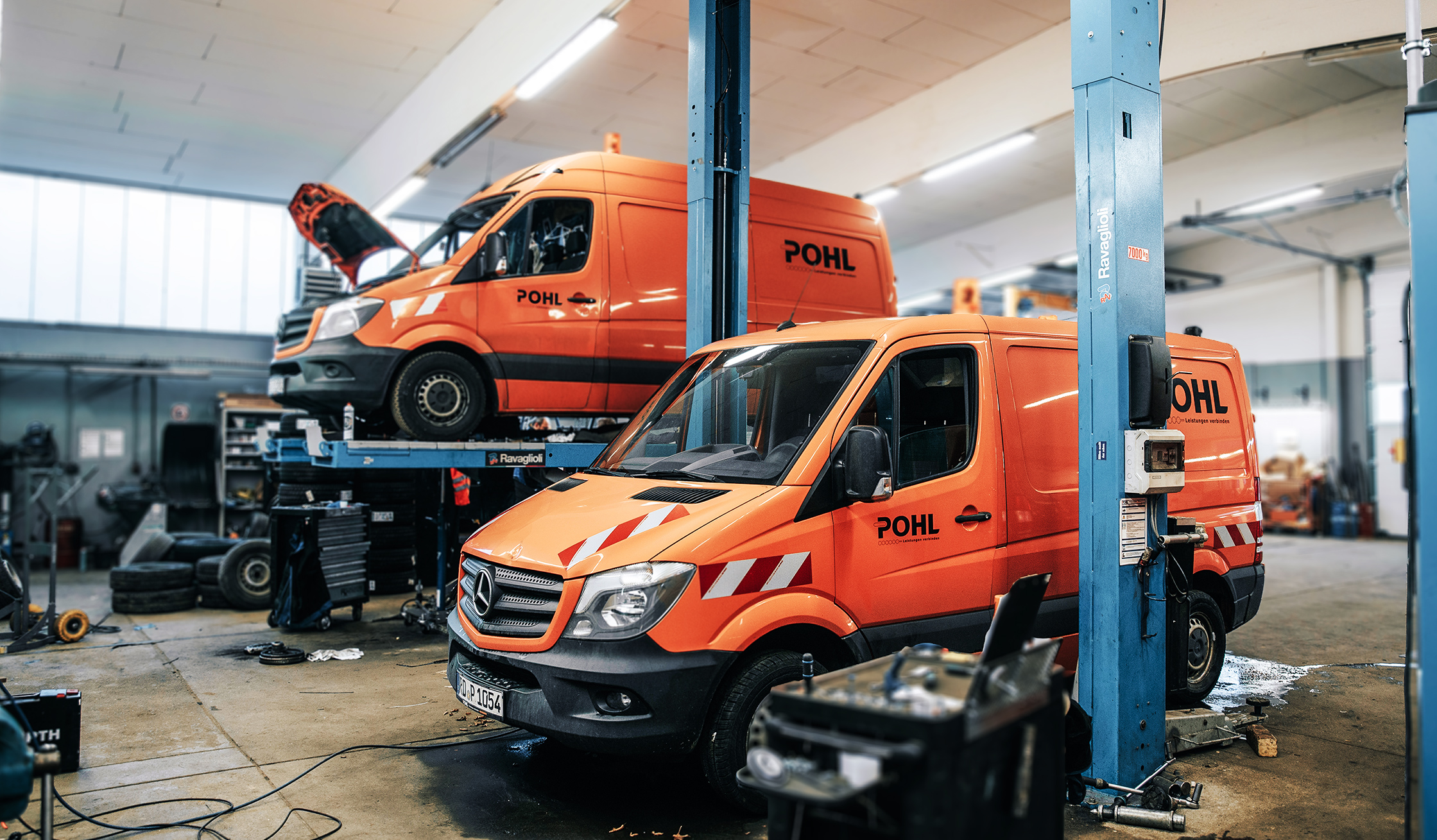 In einer Werkstatt stehen zwei orange POHL-Transporter auf Hebebühnen; Motorhauben geöffnet, daneben Werkzeugwagen und Reifen.