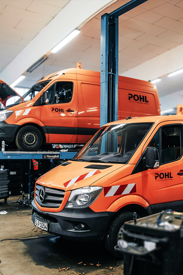 In einer Werkstatt stehen zwei orange POHL-Transporter auf Hebebühnen; Motorhauben geöffnet, daneben Werkzeugwagen und Reifen.