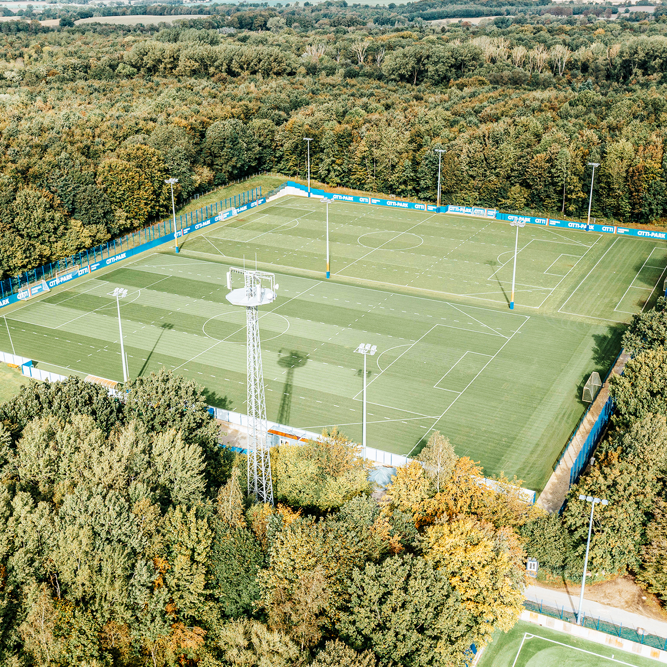 Referenz KSV Holstein Kiel Flutlicht: Ein Fußballfeld aus der Vogelperspektive, umgeben von Banden mit Werbung und Flutlichtern. Im Vordergrund Bäume und ein Funkturm.