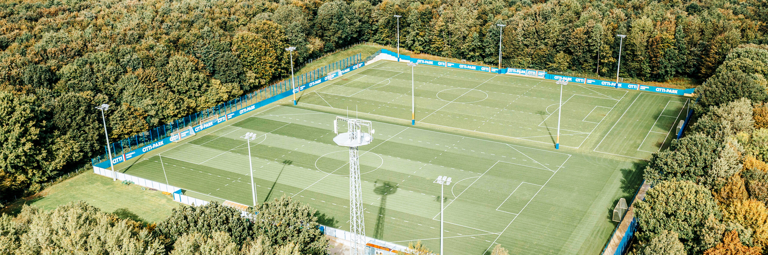 Referenz KSV Holstein Kiel Flutlicht: Ein Fußballfeld aus der Vogelperspektive, umgeben von Banden mit Werbung und Flutlichtern. Im Vordergrund Bäume und ein Funkturm.