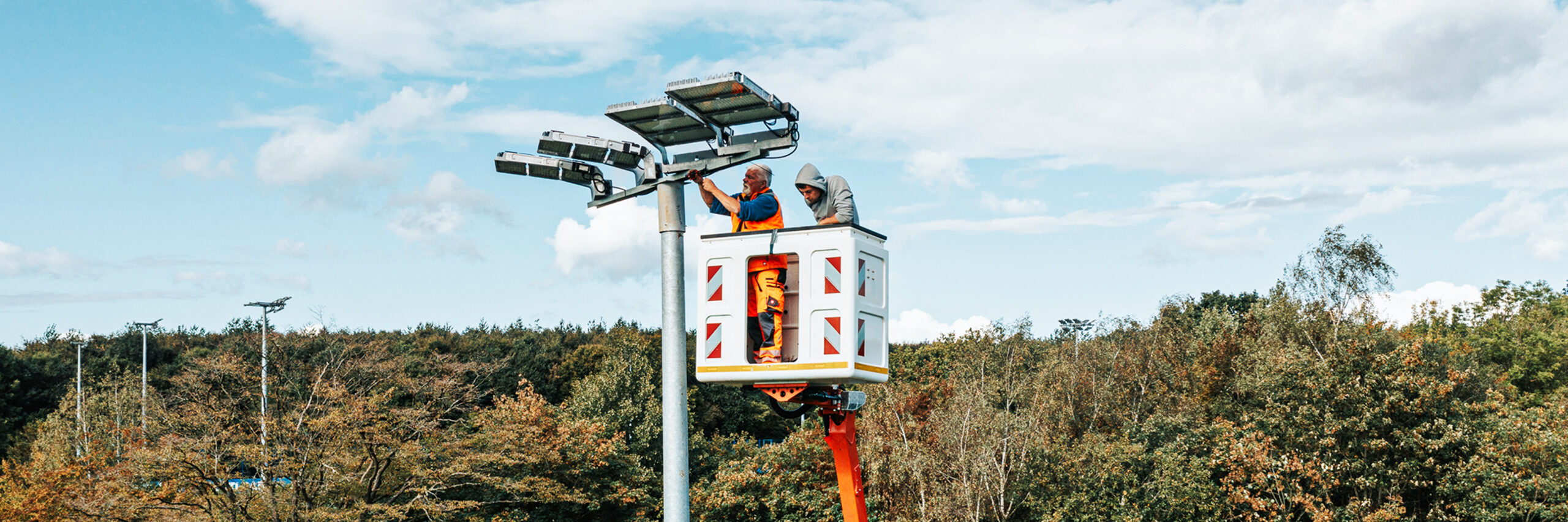 Referenz KSV Holstein Kiel Flutlicht: Oben in der Mitte arbeiten zwei POHL-Mitarbeiter im Hubkorb an mehreren großen Leuchten auf einem hohen Mast. Links unten ist der orange Ausleger der POHL-Bühne zu sehen; unten füllt dichter Baumbestand den Hintergrund.