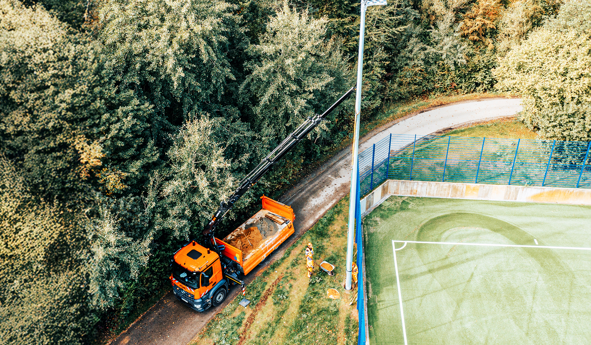 Referenz KSV Holstein Kiel Flutlicht: Unten links steht ein orangefarbener POHL-Lkw mit ausgefahrenem Kranarm auf einem schmalen Weg am Waldrand. In der Mitte ragt ein hoher Lichtmast am Rand eines Sportfelds; daneben arbeiten zwei POHL-Mitarbeiter.
