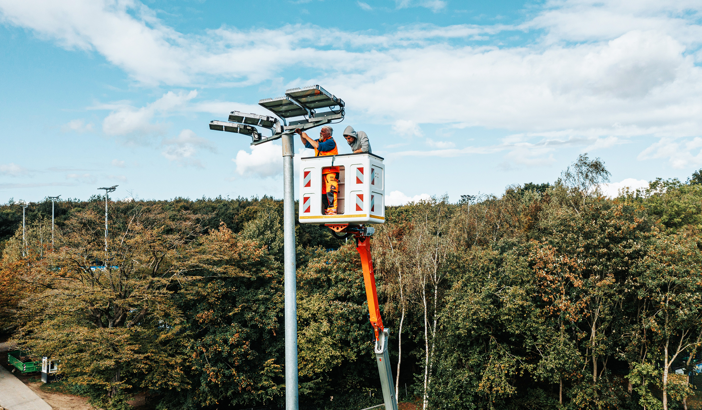 Referenz KSV Holstein Kiel Flutlicht: Oben in der Mitte arbeiten zwei POHL-Mitarbeiter im Hubkorb an mehreren großen Leuchten auf einem hohen Mast. Links unten ist der orange Ausleger der POHL-Bühne zu sehen; unten füllt dichter Baumbestand den Hintergrund.