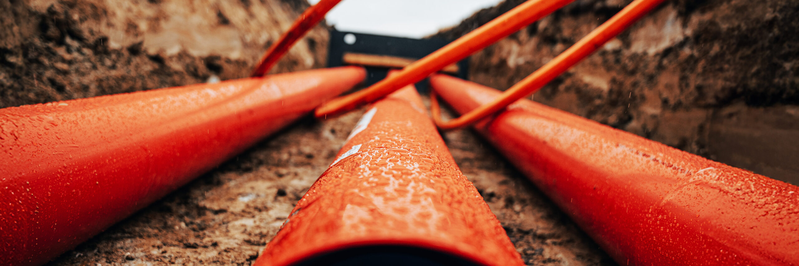 Kabel- und Leitungsbau: Mehrere orangefarbene Rohre liegen in einem aufgeschütteten Graben. Sie führen in Richtung einer Baugrube.