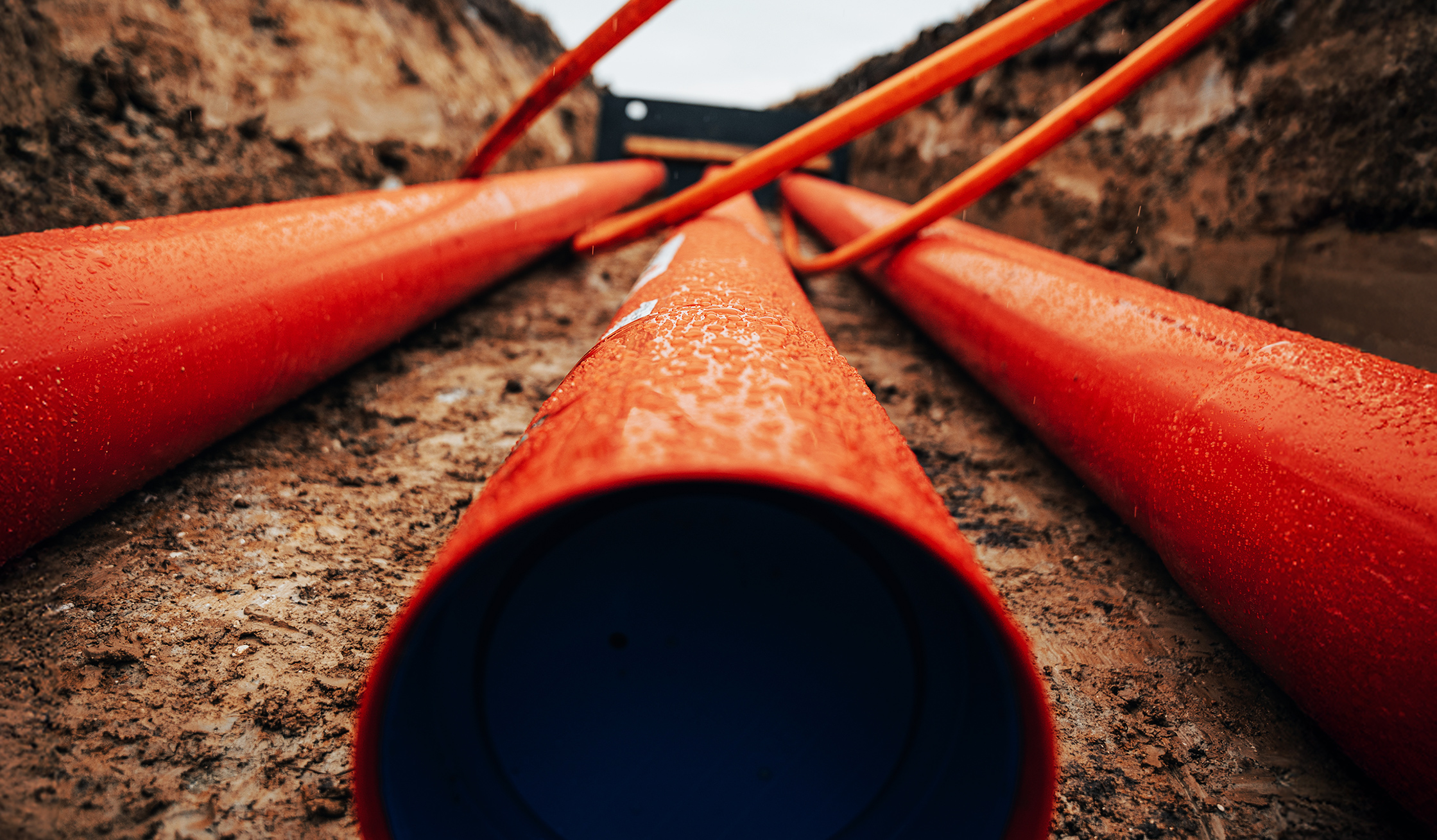 Mehrere orangefarbene Rohre liegen in einem aufgeschütteten Graben. Sie führen in Richtung einer Baugrube.
