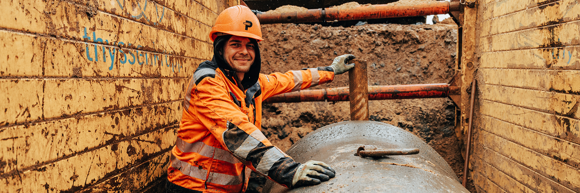 In der Mitte lehnt ein POHL-Mitarbeiter in orangefarbener Arbeitskleidung und mit orangefarbenem Helm an einem großen runden Rohr. Links und rechts im Bild stehen gelbe, erdverschmierte Wandplatten mit blauen Schriftresten. Im Hintergrund ist braune Erde zu sehen. Vorne im Bild liegt das nasse Rohr mit einem Metallhaken.