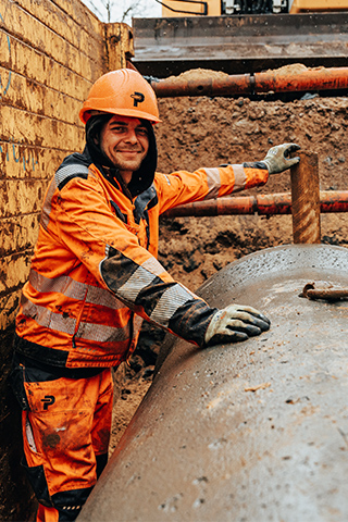 In der Mitte lehnt ein POHL-Mitarbeiter in orangefarbener Arbeitskleidung und mit orangefarbenem Helm an einem großen runden Rohr. Links und rechts im Bild stehen gelbe, erdverschmierte Wandplatten mit blauen Schriftresten. Im Hintergrund ist braune Erde zu sehen. Vorne im Bild liegt das nasse Rohr mit einem Metallhaken.