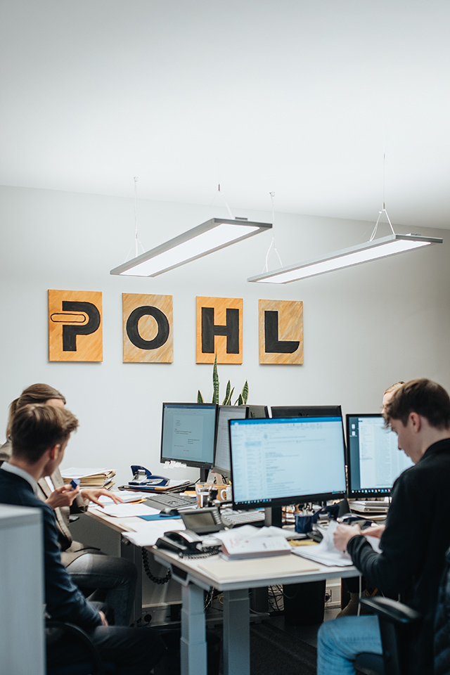 In der Mitte sitzen mehrere Personen an Schreibtischen mit Monitoren; oben an der Wand hängen vier Tafeln mit den Buchstaben „POHL“, rechts im Raum stehen helle Schränke.