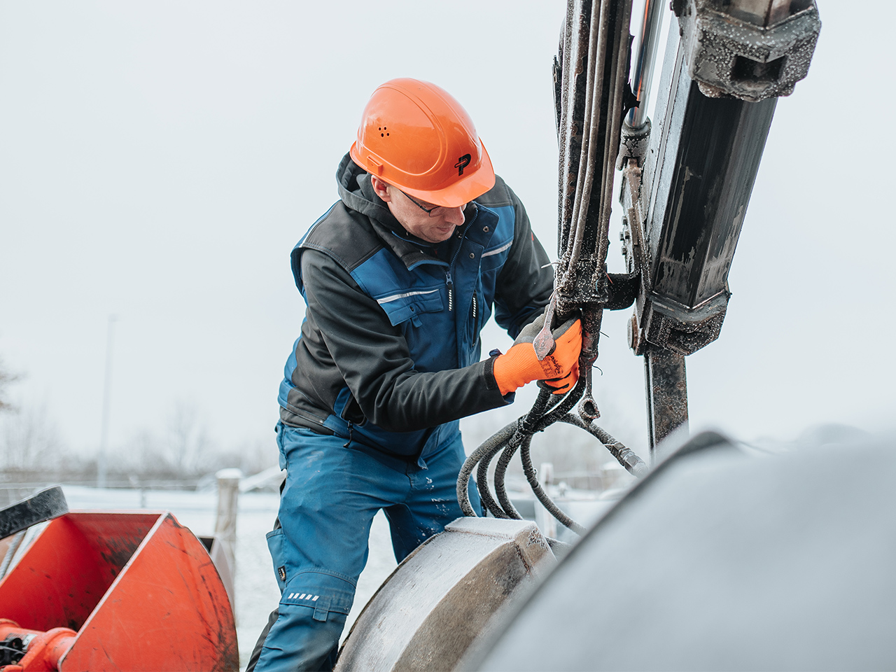 In der Mitte arbeitet ein POHL-Mitarbeiter mit orangefarbenem Helm und Handschuhen an dicken Schläuchen an einem großen Maschinenarm; links im Bild steht eine rote Anbauschaufel, im Hintergrund ist grauer Himmel.