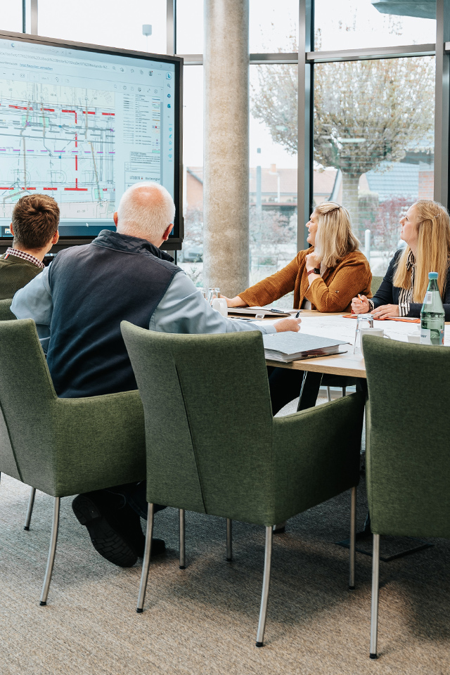 In der Mitte sitzen sechs Personen an einem runden Tisch und schauen auf einen großen Bildschirm links im Bild mit Plänen; auf dem Tisch stehen Tassen mit POHL-Logo, im Hintergrund sind große Fenster mit Blick nach draußen.