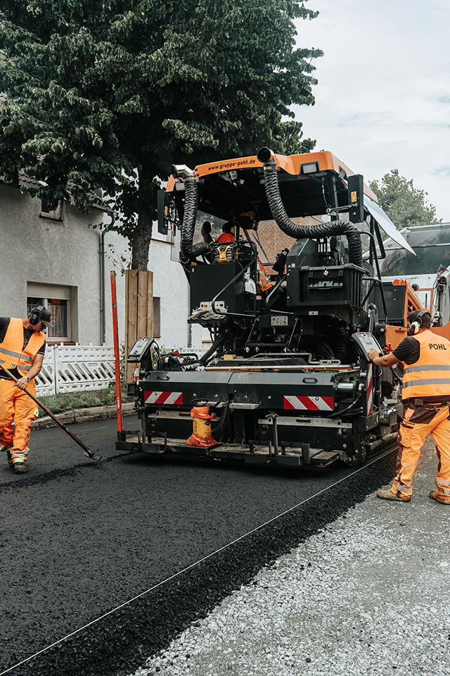 In der Mitte arbeitet eine große Straßenbaumaschine auf frischem Asphalt; links und rechts stehen POHL-Mitarbeiter in orangefarbener Kleidung mit Werkzeugen; im Hintergrund fährt eine Reihe orangefarbener POHL-Fahrzeuge die Straße entlang.