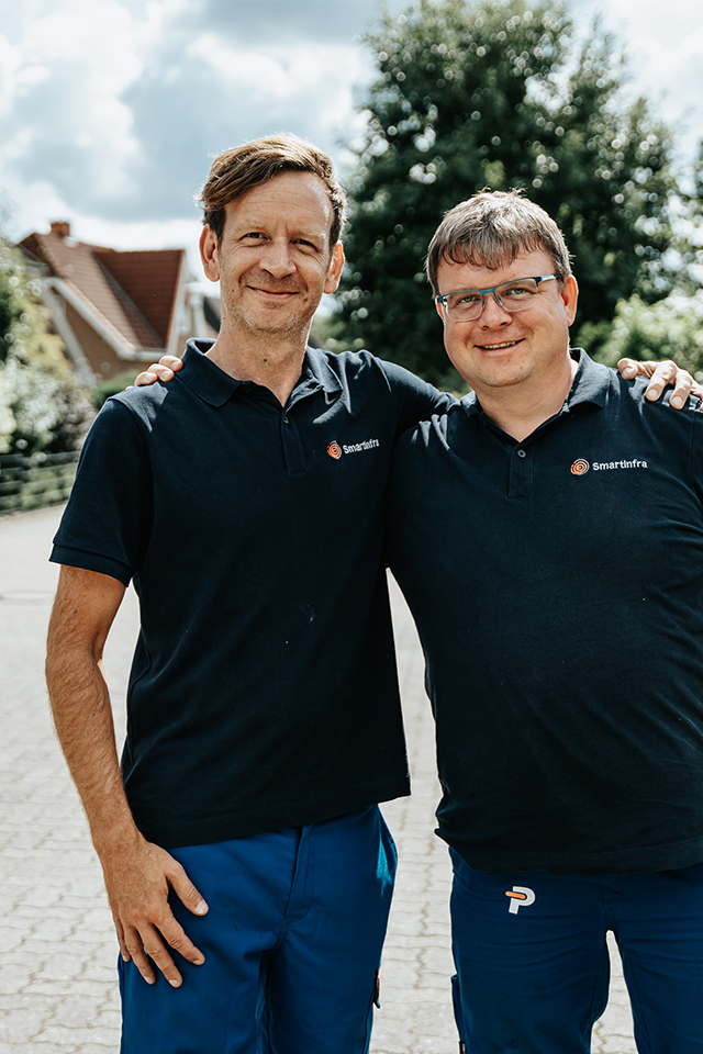 Auf dem Bild stehen zwei Männer, die Arm in Arm freundlich in die Kamera lächeln. Sie haben ein schwarzes T-Shirt an. Auf dem T-Shirt ist oben rechts das Smartinfra Logo der POHL-Gruppe. Im Hintergrund ist ein Weg mit grünen Bäumen.