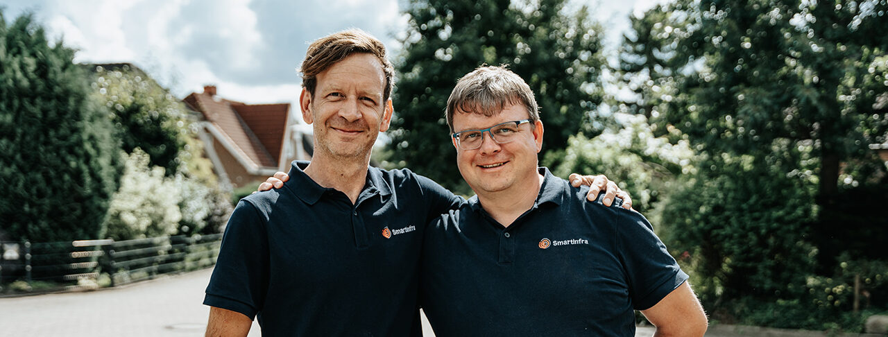 Auf dem Bild stehen zwei Männer, die Arm in Arm freundlich in die Kamera lächeln. Sie haben ein schwarzes T-Shirt an. Auf dem T-Shirt ist oben rechts das Smartinfra Logo der POHL-Gruppe. Im Hintergrund ist ein Weg mit grünen Bäumen.