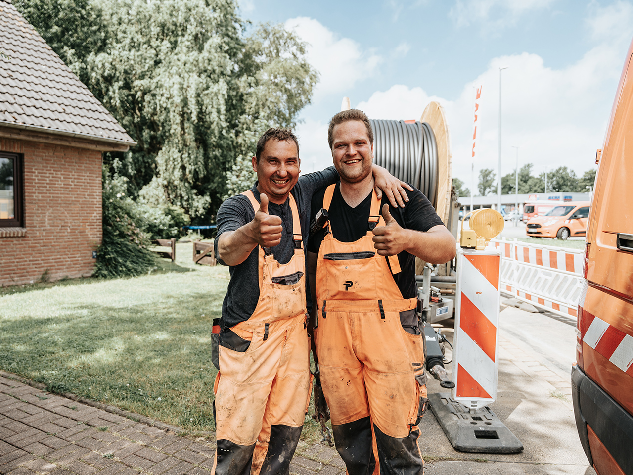 In der Mitte stehen zwei POHL-Mitarbeiter in orangefarbenen Latzhosen nebeneinander; sie zeigen den Daumen nach oben, hinter ihnen liegen eine große Kabeltrommel und weiß-orange Absperrgitter, rechts am Rand steht ein orangefarbener POHL-Transporter.