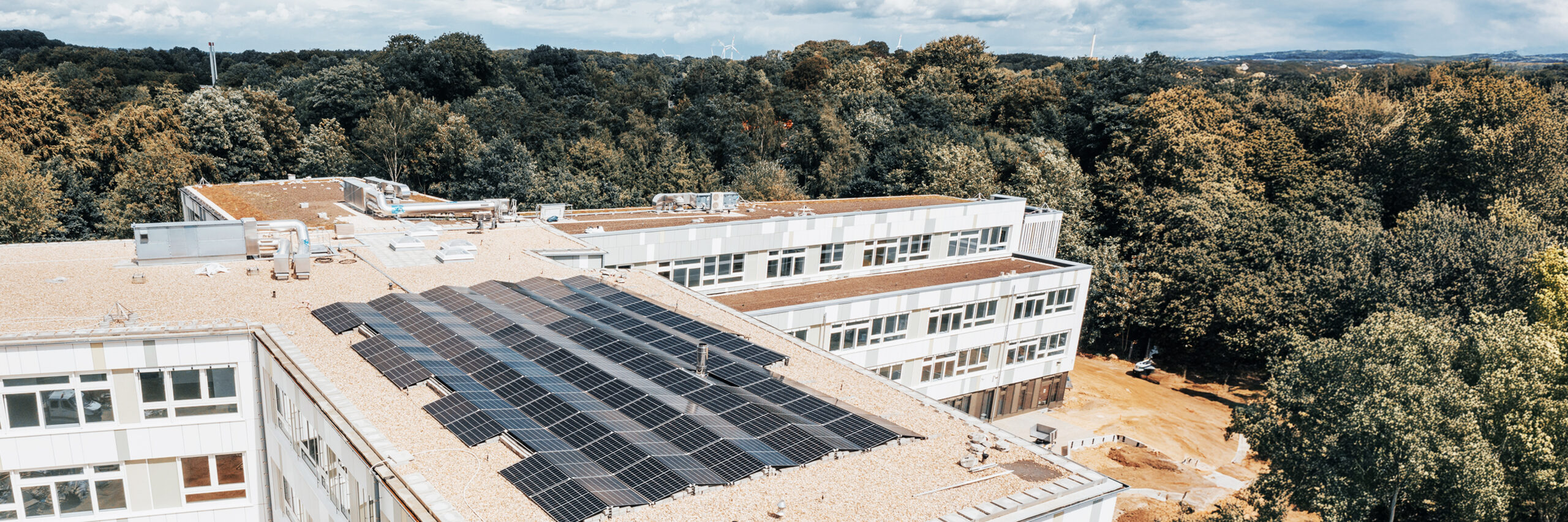 Referenz Elektrotechnik Schule Kellinghusen: In der Mitte bedecken in mehreren Reihen schwarze Solarmodule das flache Kiesdach eines großen Gebäudes. Im Hintergrund schließen weitere Gebäudeteile an; dahinter beginnt ein breiter Gürtel aus Bäumen.