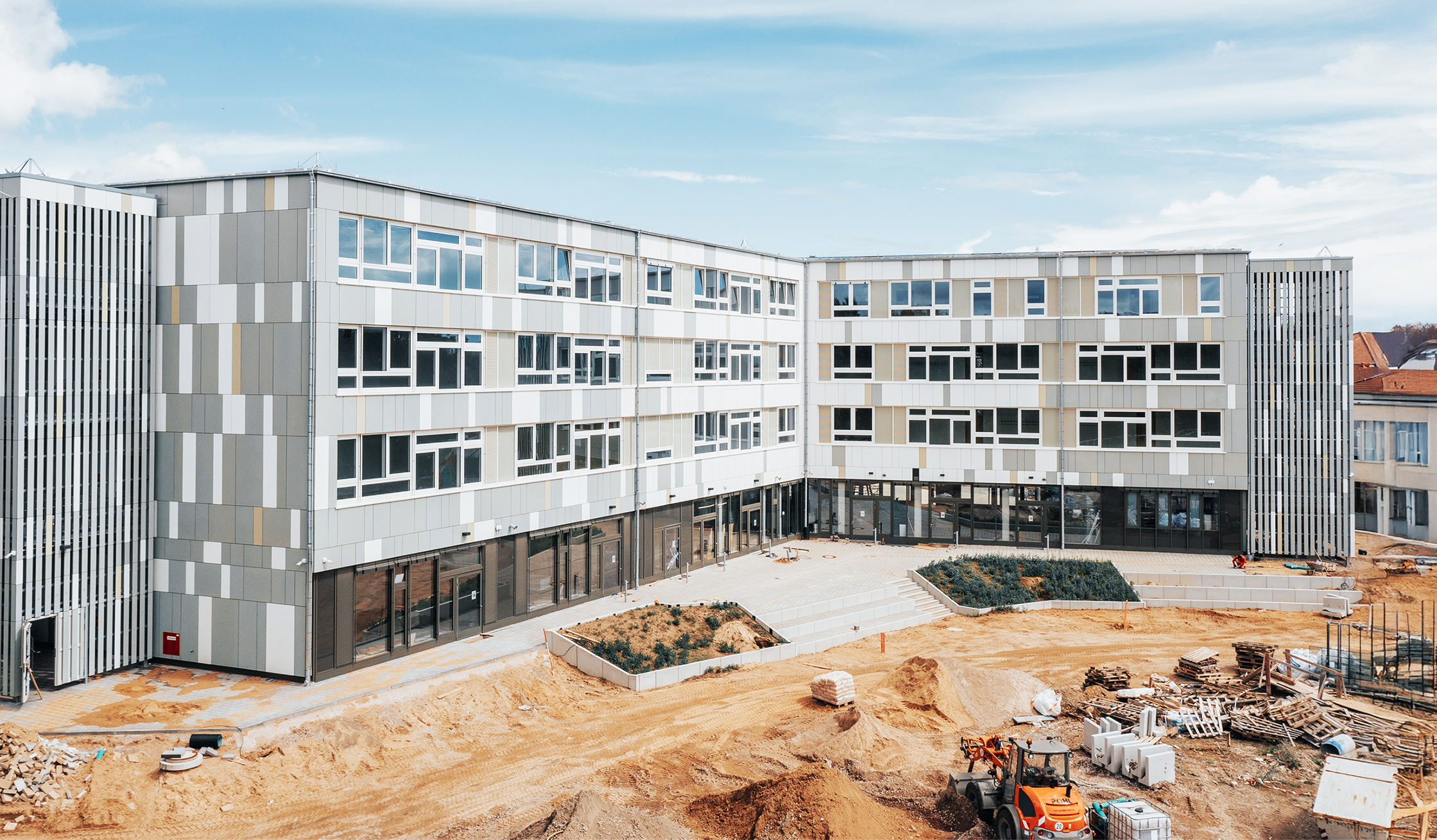 Referenz Elektrotechnik Schule Kellinghusen: Vor einem U-förmigen Gebäude liegt eine offene Baustelle. Unten rechts steht eine orange POHL-Maschine mit Schaufel neben Erdhaufen und gestapelten Materialien. In der Mitte des Hofs liegen zwei eingefasste Beete mit Treppen; oben über der Fassade ist blauer Himmel.