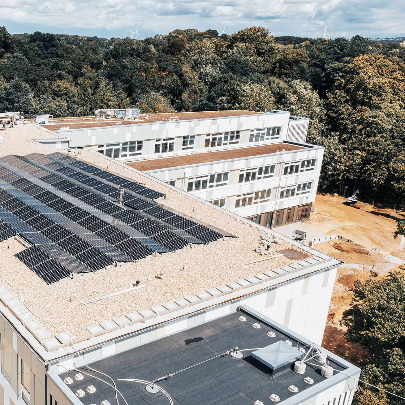 Referenz Elektrotechnik Schule Kellinghusen: In der Mitte bedecken in mehreren Reihen schwarze Solarmodule das flache Kiesdach eines großen Gebäudes. Im Hintergrund schließen weitere Gebäudeteile an; dahinter beginnt ein breiter Gürtel aus Bäumen.