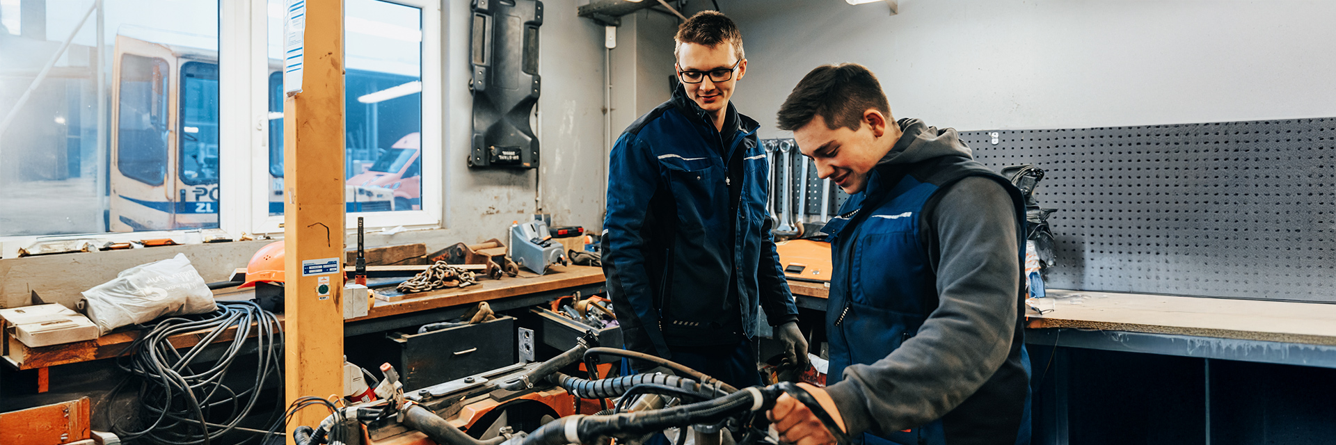 In der Mitte arbeiten zwei Personen in dunkelblauen Westen an einem Gerät auf einer Werkbank. Links im Bild steht ein Fenster; dahinter ist ein orangefarbenes Fahrzeug mit sichtbarem POHL-Schriftzug zu sehen. Auf der Werkbank liegen ein orangefarbener Helm, Schläuche und Werkzeuge. Rechts im Bild hängt eine dunkle Lochwand über der Arbeitsfläche. Im Hintergrund stehen Regale und weitere Werkstattteile.