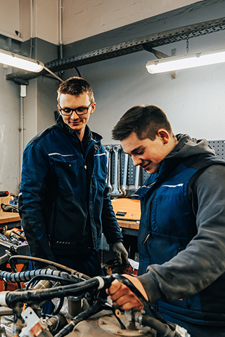 In der Mitte arbeiten zwei Personen in dunkelblauen Westen an einem Gerät auf einer Werkbank. Links im Bild steht ein Fenster; dahinter ist ein orangefarbenes Fahrzeug mit sichtbarem POHL-Schriftzug zu sehen. Auf der Werkbank liegen ein orangefarbener Helm, Schläuche und Werkzeuge. Rechts im Bild hängt eine dunkle Lochwand über der Arbeitsfläche. Im Hintergrund stehen Regale und weitere Werkstattteile.