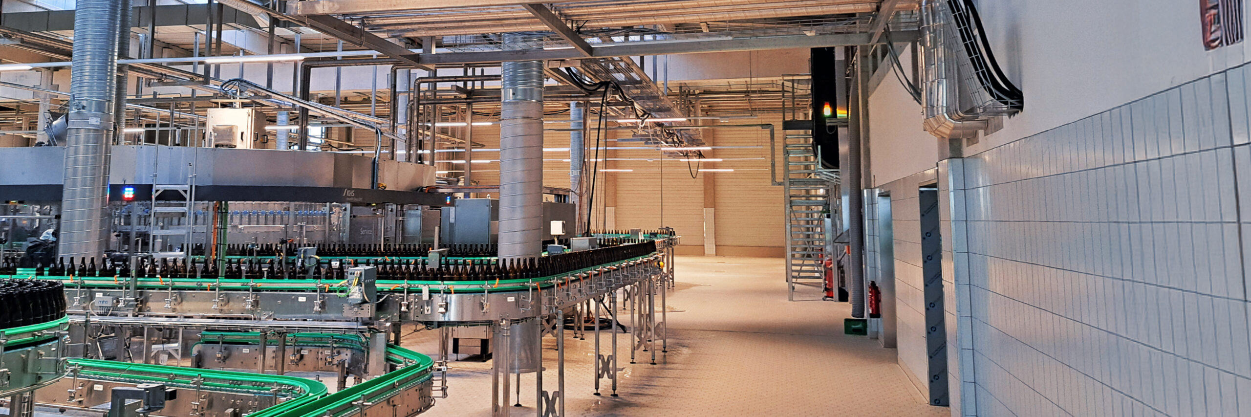 Referenz Marne Dithmarscher Brauerei: Innenansicht einer modernen Produktionshalle mit Förderbändern voller Flaschen und zahlreichen Rohren und Leitungen unter der Hallendecke.