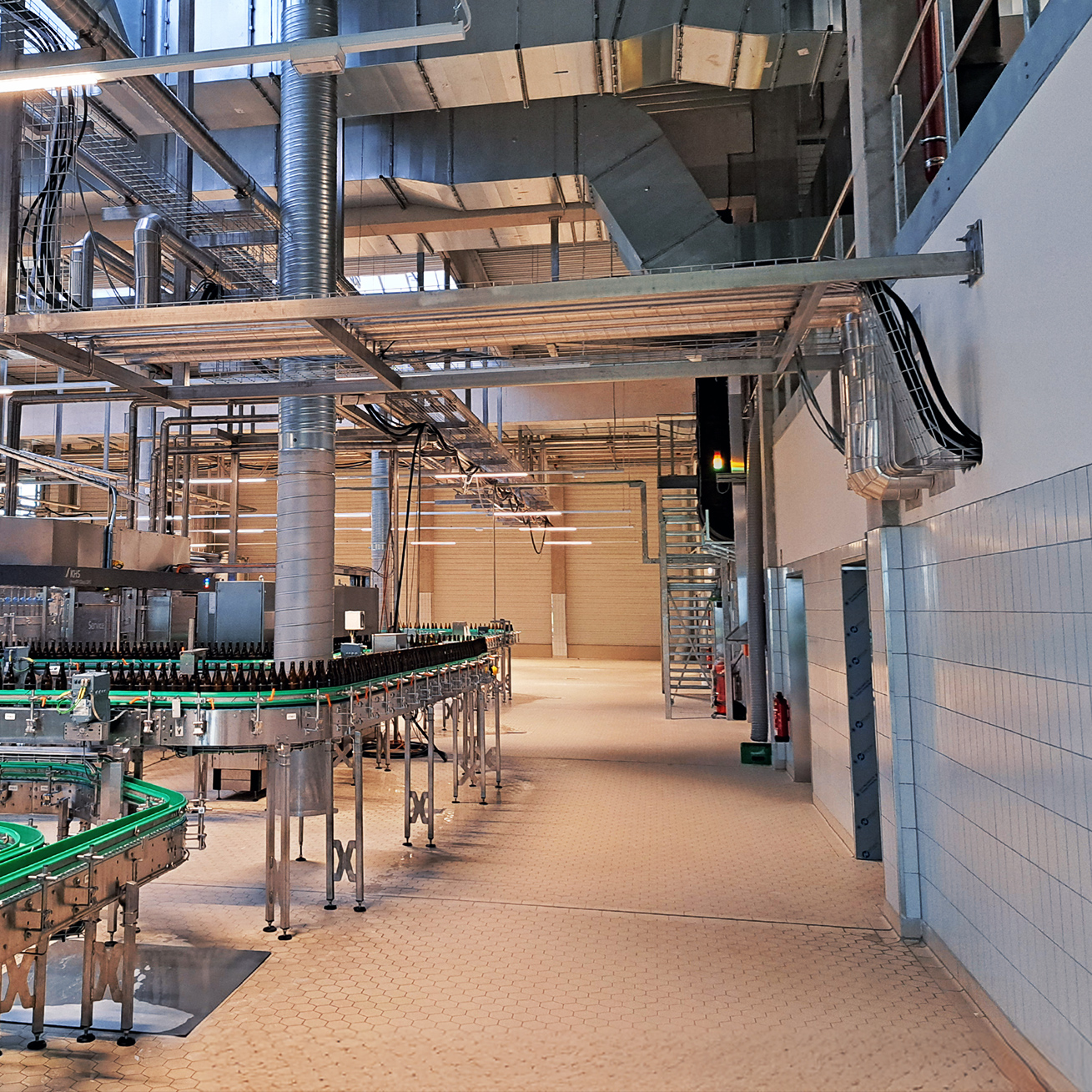 Referenz Marne Dithmarscher Brauerei: Innenansicht einer modernen Produktionshalle mit Förderbändern voller Flaschen und zahlreichen Rohren und Leitungen unter der Hallendecke.