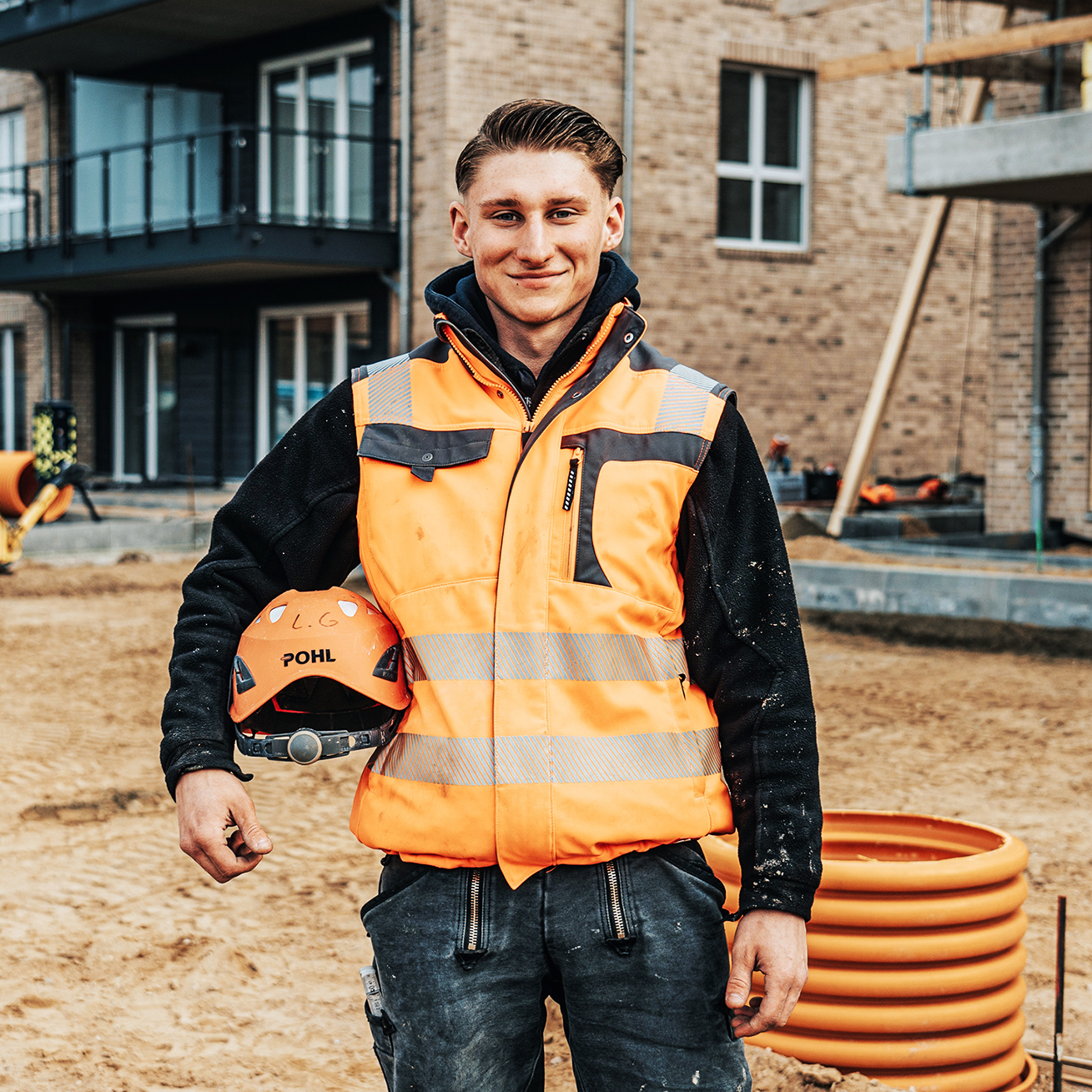Ein POHL-Mitarbeiter in orangefarbener Weste steht auf einer Baustelle vor einem Neubau mit Balkonen; er hält einen Helm mit POHL-Logo, rechts liegt eine orange Rohrspirale auf sandigem Boden.