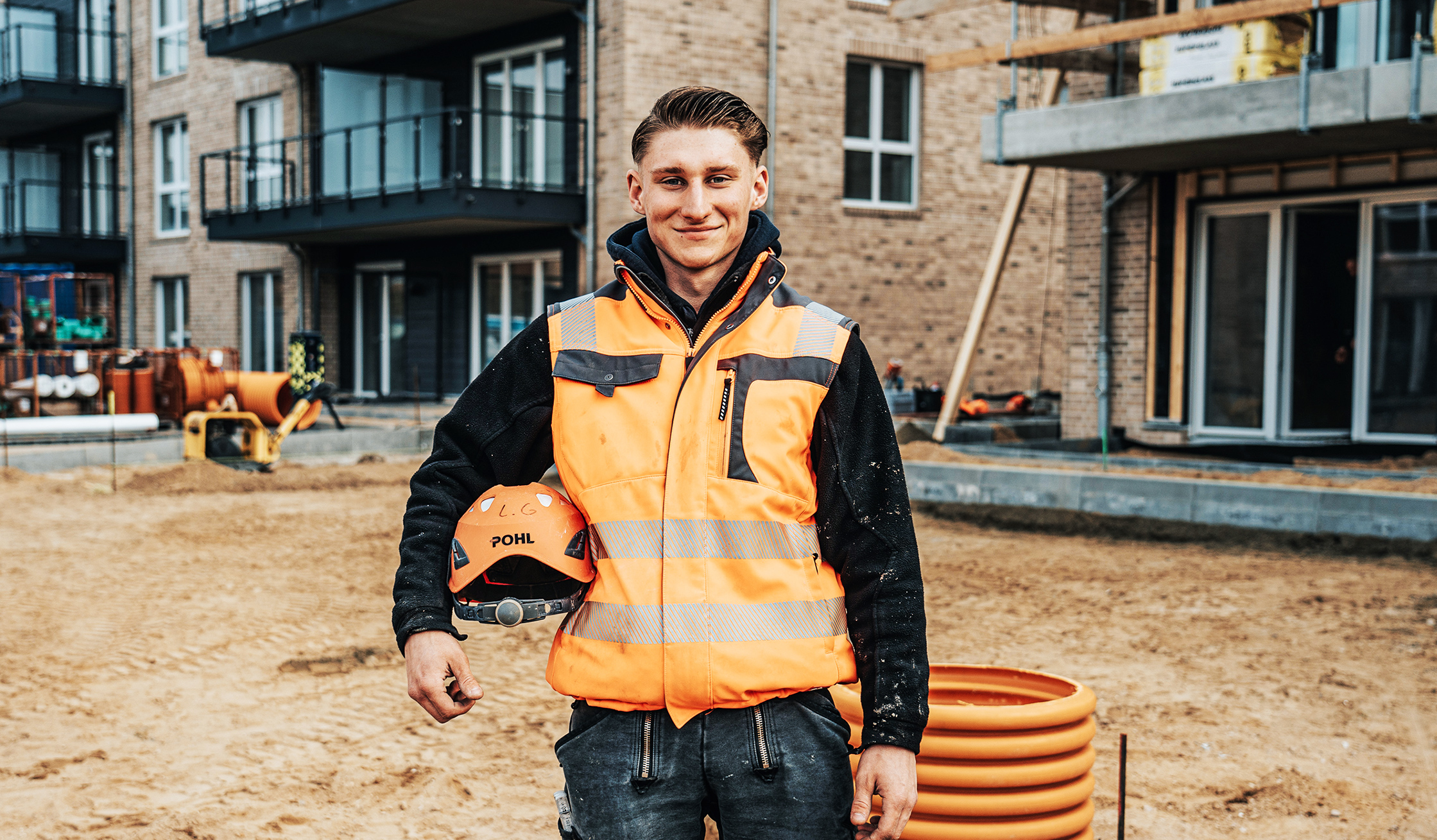 Ein POHL-Mitarbeiter in orangefarbener Weste steht auf einer Baustelle vor einem Neubau mit Balkonen; er hält einen Helm mit POHL-Logo, rechts liegt eine orange Rohrspirale auf sandigem Boden.