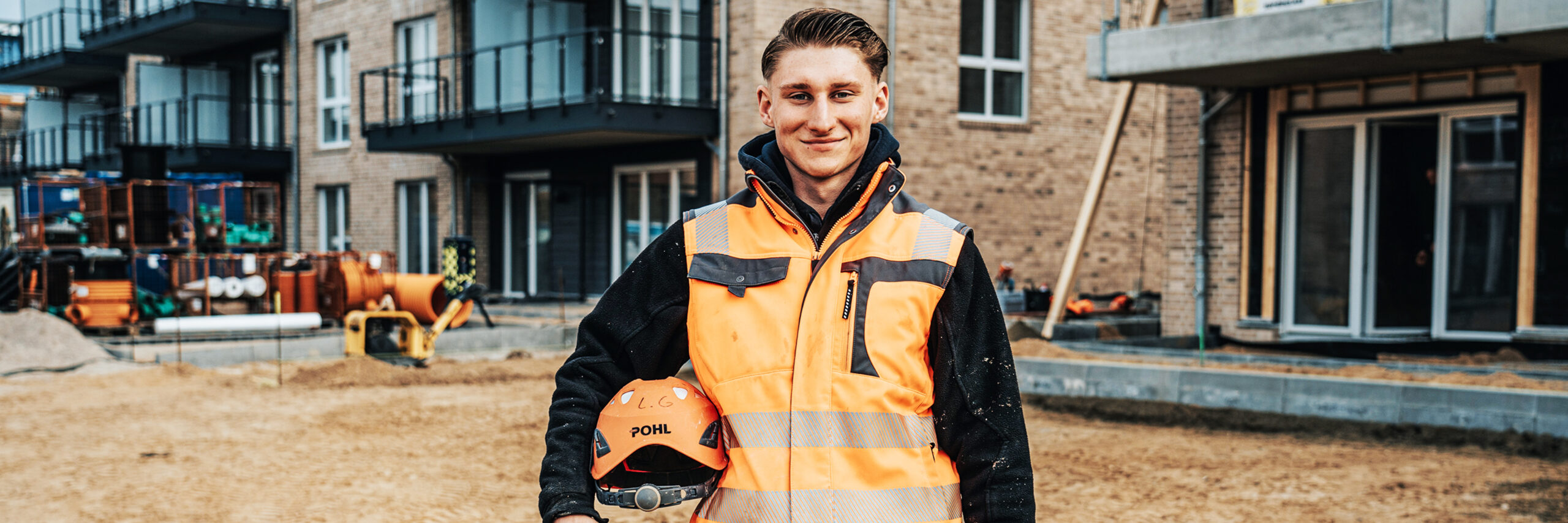 Ein POHL-Mitarbeiter in orangefarbener Weste steht auf einer Baustelle vor einem Neubau mit Balkonen; er hält einen Helm mit POHL-Logo, rechts liegt eine orange Rohrspirale auf sandigem Boden.
