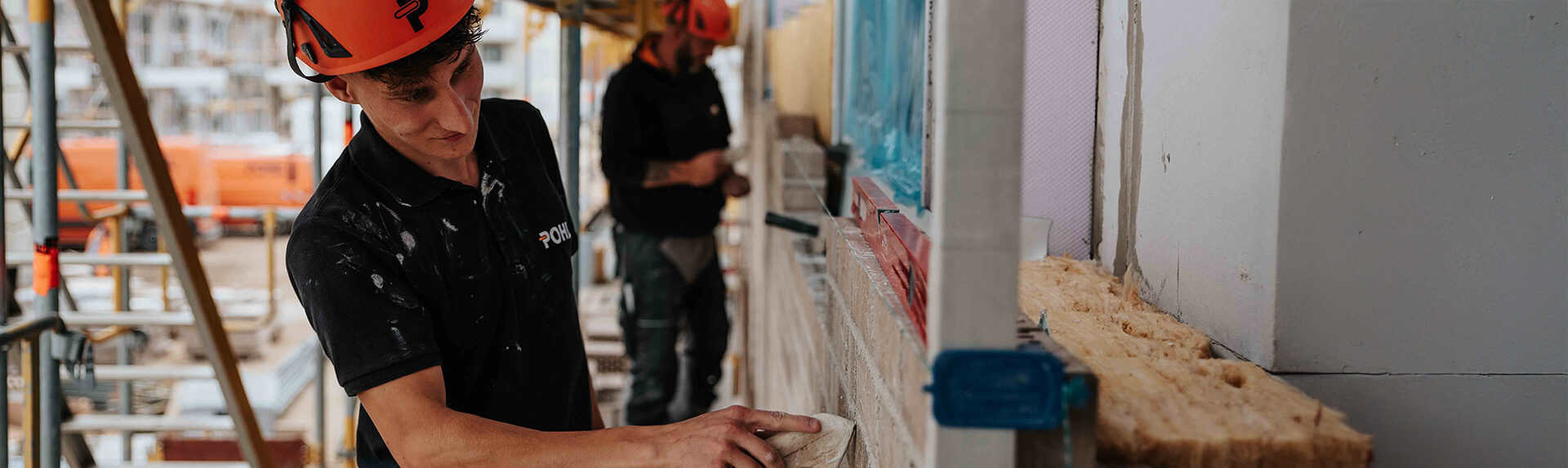 In der Mitte trägt ein POHL-Mitarbeiter mit orangefarbenem Helm Mörtel auf eine Mauer auf; rechts im Bild liegt gelbes Dämmmaterial, im Hintergrund steht ein Gerüst mit weiterem POHL-Mitarbeiter.