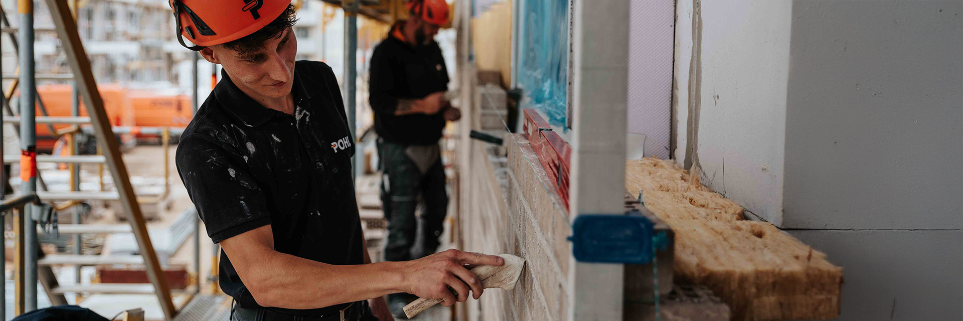 In der Mitte trägt ein POHL-Mitarbeiter mit orangefarbenem Helm Mörtel auf eine Mauer auf; rechts im Bild liegt gelbes Dämmmaterial, im Hintergrund steht ein Gerüst mit weiterem POHL-Mitarbeiter.