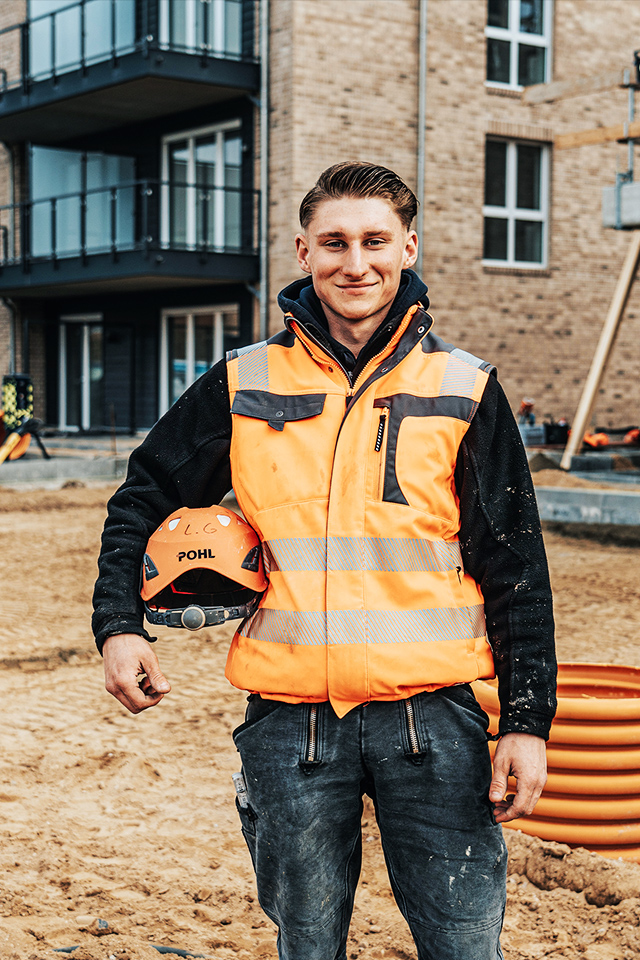 Ein POHL-Mitarbeiter in orangefarbener Weste steht auf einer Baustelle vor einem Neubau mit Balkonen; er hält einen Helm mit POHL-Logo, rechts liegt eine orange Rohrspirale auf sandigem Boden.