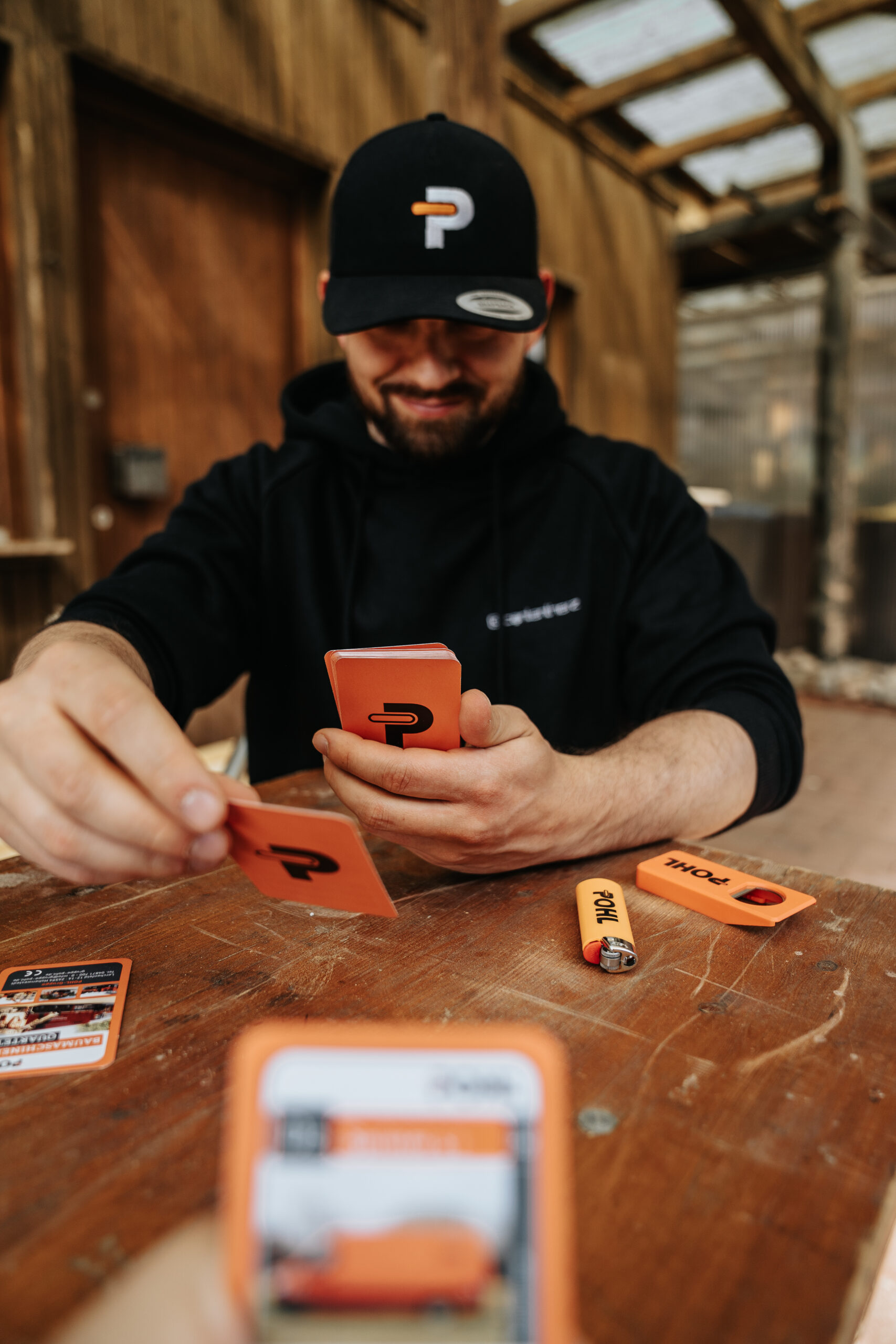 Ein Mann sitzt an einem Holztisch, hält orange Spielkarten in der Hand und legt eine Karte ab, auf dem Tisch liegen ein Feuerzeug und ein Flaschenöffner mit Pohl Schriftzug, der Mann trägt eine schwarze Kappe mit Pohl Logo und einen Hoodie, im Hintergrund steht eine hölzerne Überdachung, die Szene wirkt entspannt.