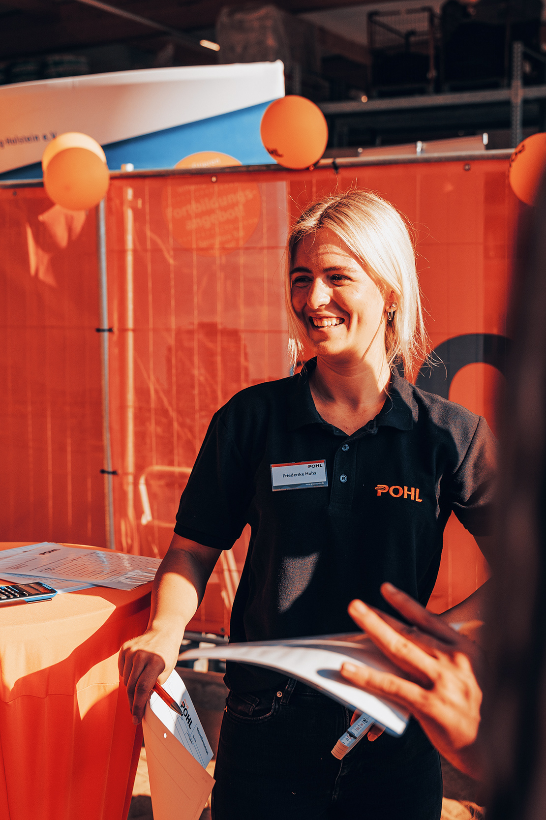 Eine Frau im schwarzen Pohl-Poloshirt steht an einem Infotisch mit Unterlagen, über ihr hängen orange Luftballons, im Hintergrund ein orangefarbener Messe-Backdrop.