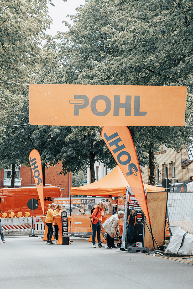 Über der Straße hängt ein großes Pohl-Banner, darunter ein orangefarbenes Zelt mit zwei Pohl-Flaggen links und rechts sowie mehreren Personen vor Infoflächen.