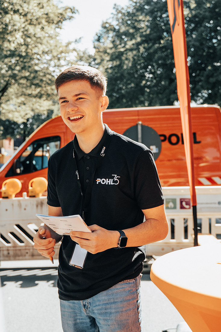 Ein junger Mann im schwarzen Pohl-Poloshirt lächelt im Vordergrund und hält ein Klemmbrett, hinter ihm stehen eine Pohl-Flagge, eine Absperrung und ein orangefarbener Transporter.