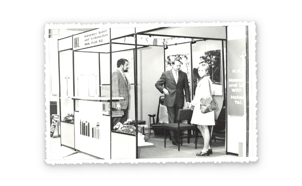 Historische Messeaufnahme in Schwarzweiß, drei Erwachsene am offenen Messestand mit filigranen Rahmen, im Hintergrund Schilder mit Pohl Schriftzug.