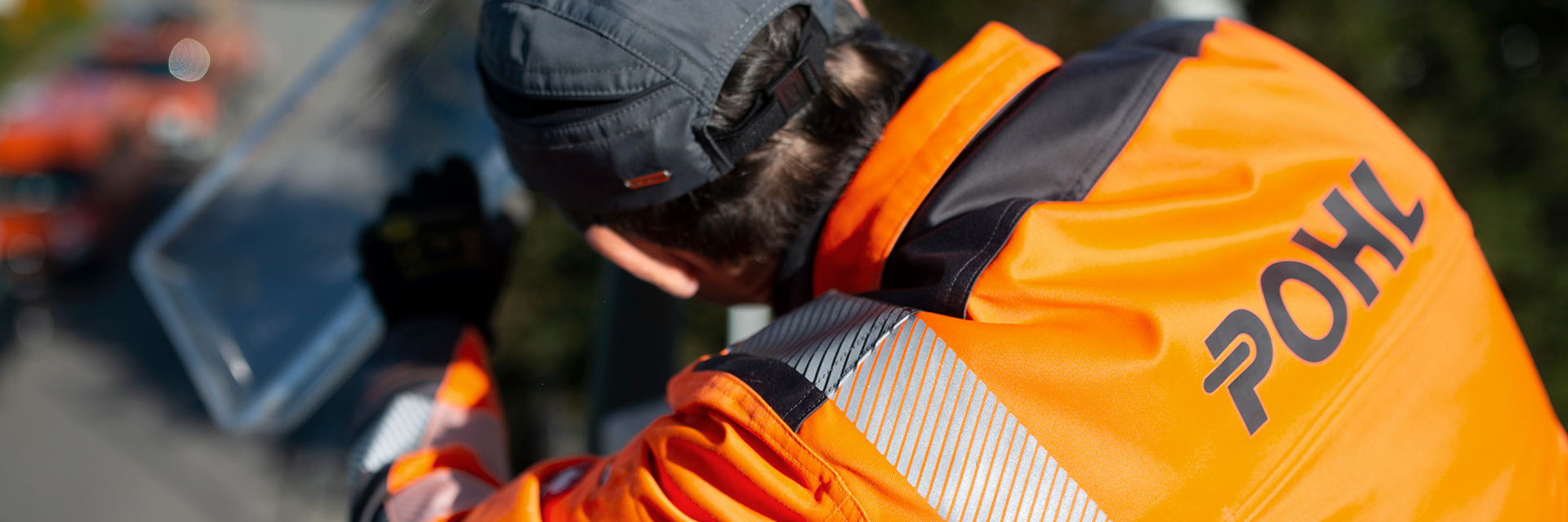 Vorne im Bild steht ein POHL-Mitarbeiter in orangefarbener Jacke von hinten und arbeitet links oben an einer Leuchte; im Hintergrund sind Bäume und eine Straße.