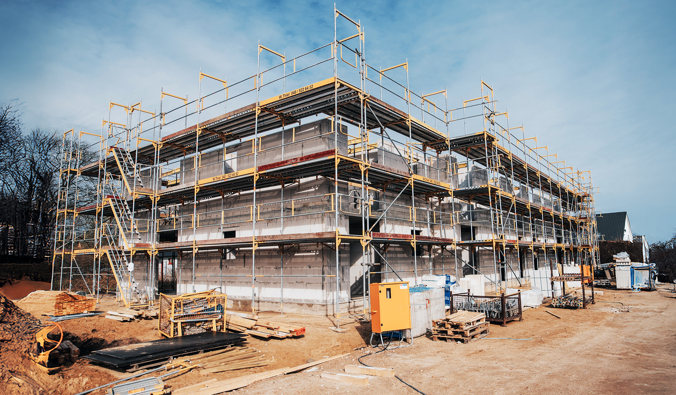 Referenz Mehrfamilienhäuser Neumünster: Eingerüsteter Rohbau eines mehrstöckigen Gebäudes mit Baumaterialien im Vordergrund