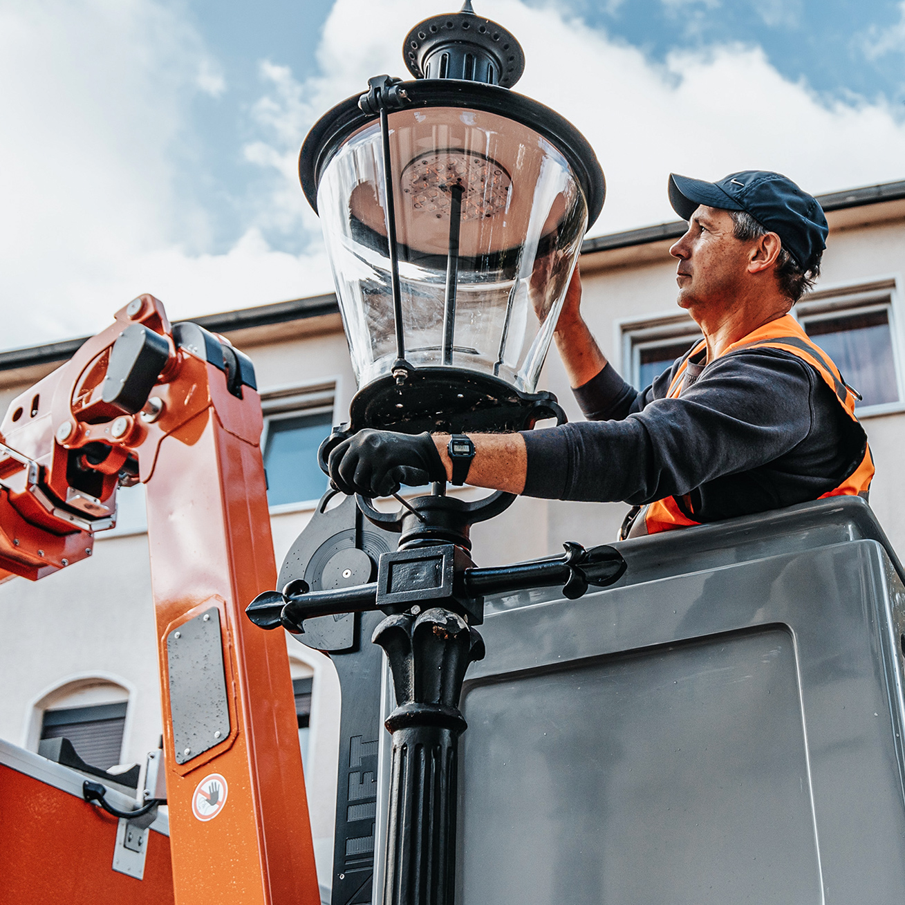 Referenz Wartung Laternen Neumünster: In der Mitte steht ein POHL-Mitarbeiter im Arbeitskorb und fasst an den Glasaufsatz einer Straßenlaterne. Links ragt der orange Ausleger der Bühne ins Bild; hinten Hausfassade.