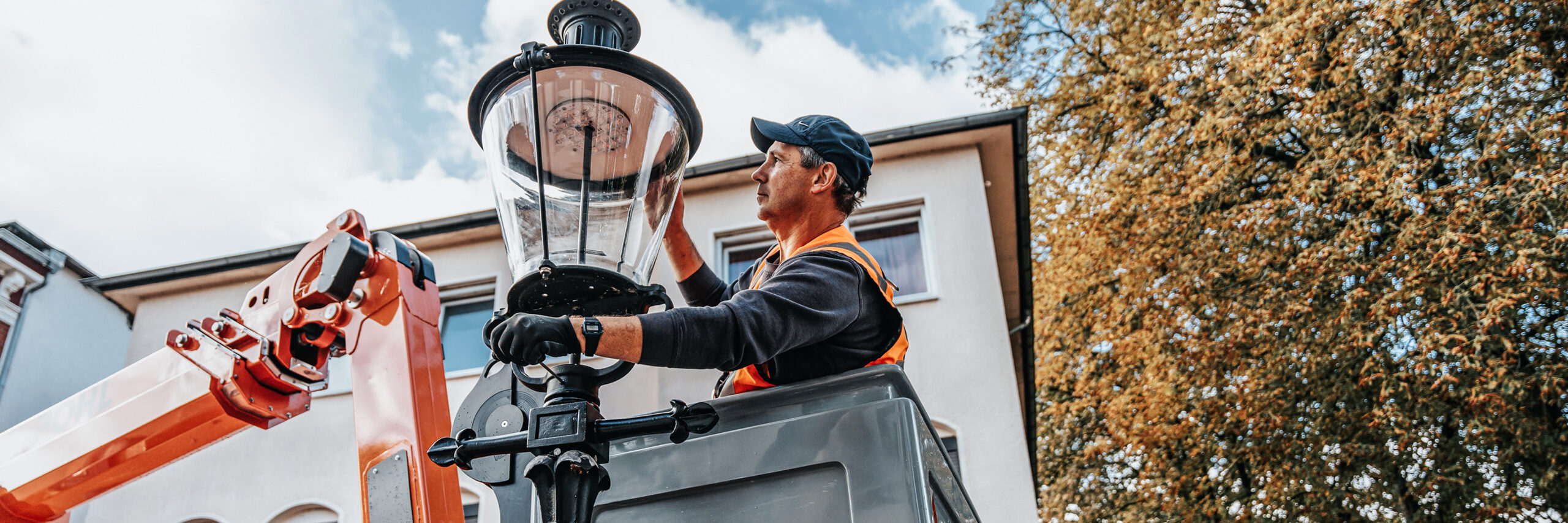 Referenz Wartung Laternen Neumünster: In der Mitte steht ein POHL-Mitarbeiter im Arbeitskorb und fasst an den Glasaufsatz einer Straßenlaterne. Links ragt der orange Ausleger der Bühne ins Bild; hinten Hausfassade, rechts Baumkrone.