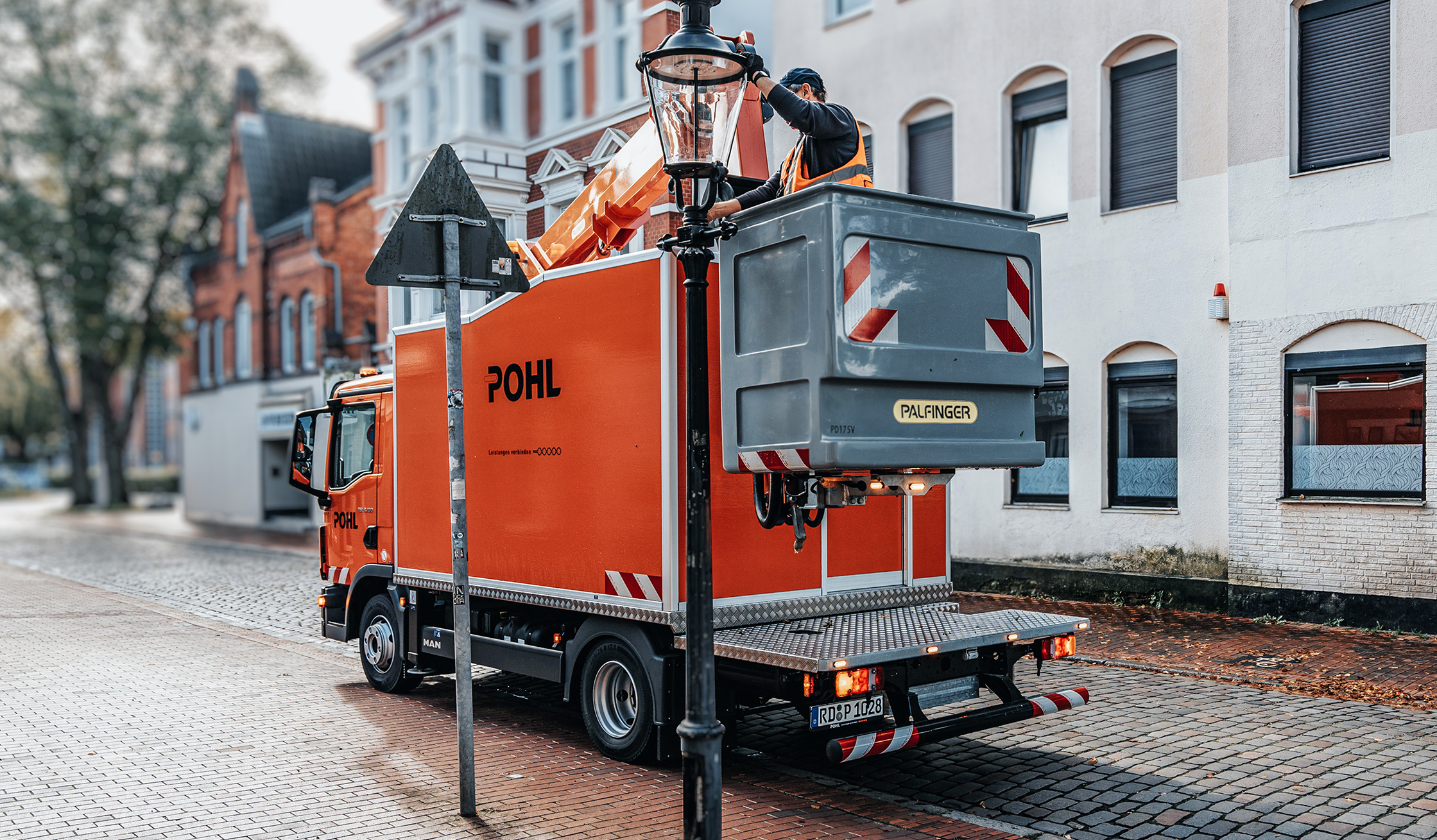 Referenz Wartung Laternen Neumünster: Links parkt ein orangefarbener POHL-Lkw mit Hubkorb. In der Mitte arbeitet ein POHL-Mitarbeiter an einer Laterne; rechts eine helle Hausfassade mit Fenstern.