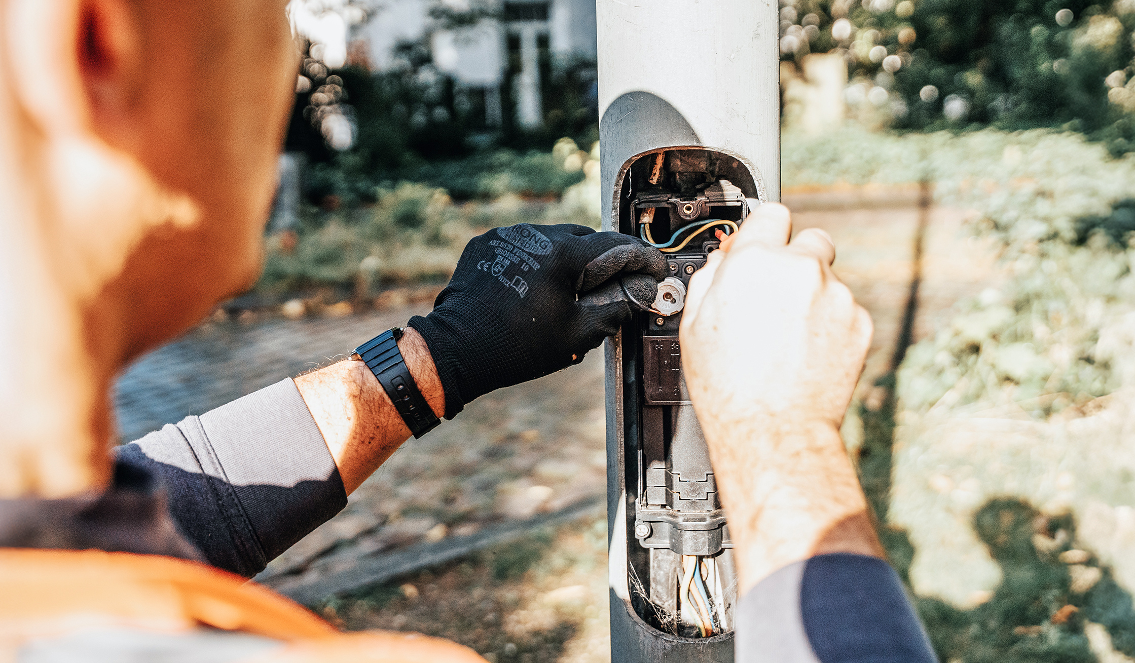 Referenz Wartung Laternen Neumünster: Vorne links greifen behandschuhte Hände eines POHL-Mitarbeiters in den geöffneten Fuß einer Straßenlaterne. In der Mitte sind Kabel und Bauteile zu sehen; im Hintergrund Gehweg und Grün unscharf.