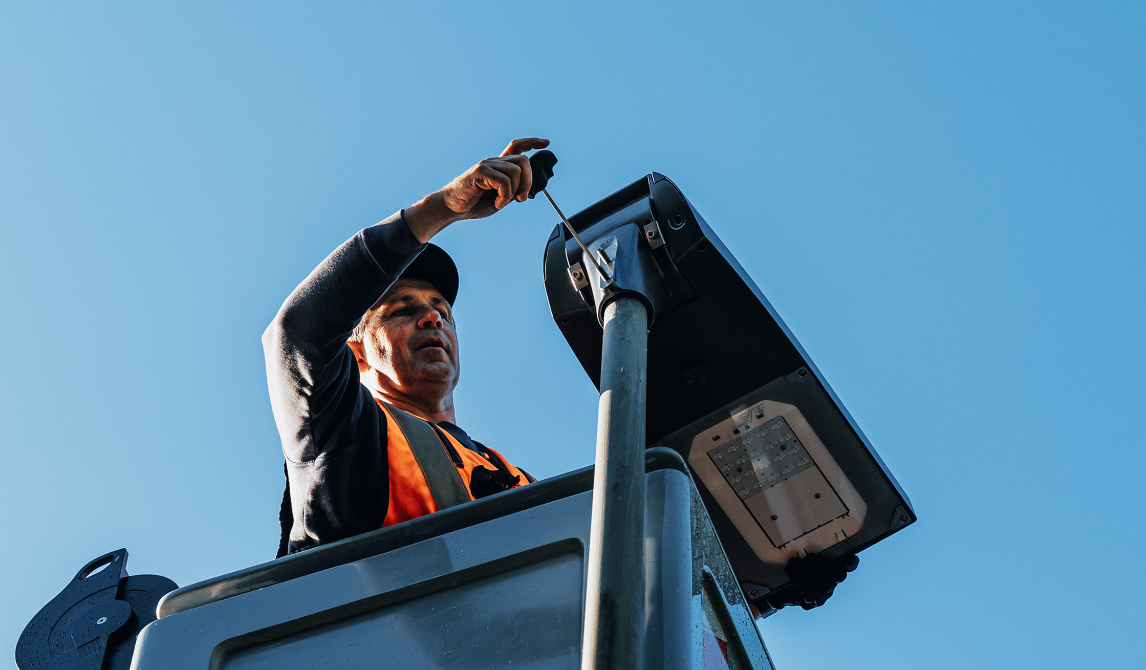Referenz Umrüstung LED Neumünster: Oben in der Mitte justiert ein POHL-Mitarbeiter im Korb den Lampenkopf an einem Mast. Links ist der Korbrand zu sehen, rechts liegt freier Himmel.