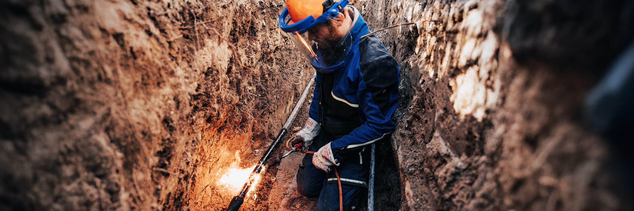 Energieinfrastruktur: Ein POHL Arbeiter mit orangefarbenem Helm und Gesichtsschutz befindet sich in einem tiefen Graben. Er verschweißt ein Kabel, Funken und Flammen sind sichtbar.