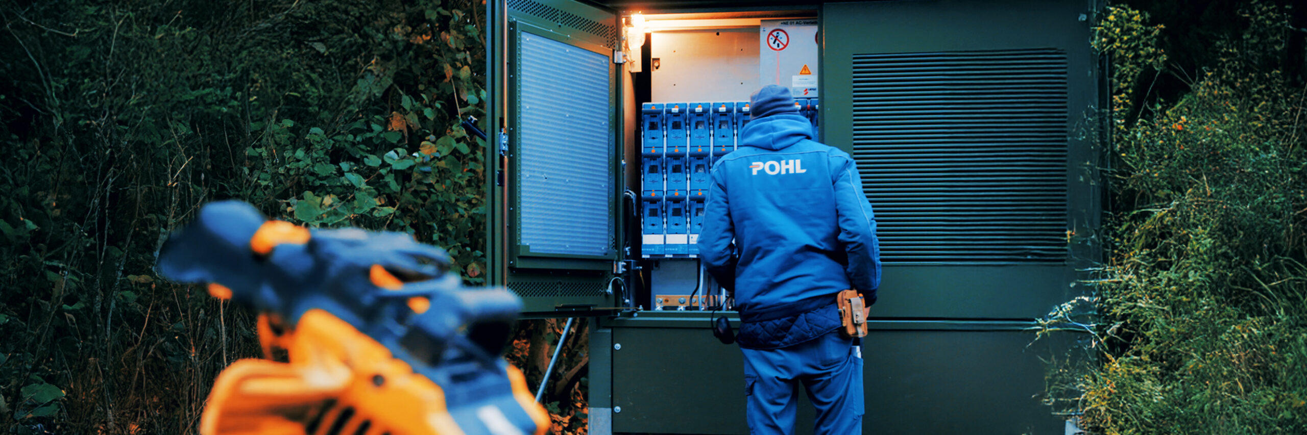 Ein Monteur in blauer POHL-Jacke arbeitet an einem großen grünen Schaltschrank im Freien. Das Innere mit vielen Schaltern und Kabeln ist beleuchtet.