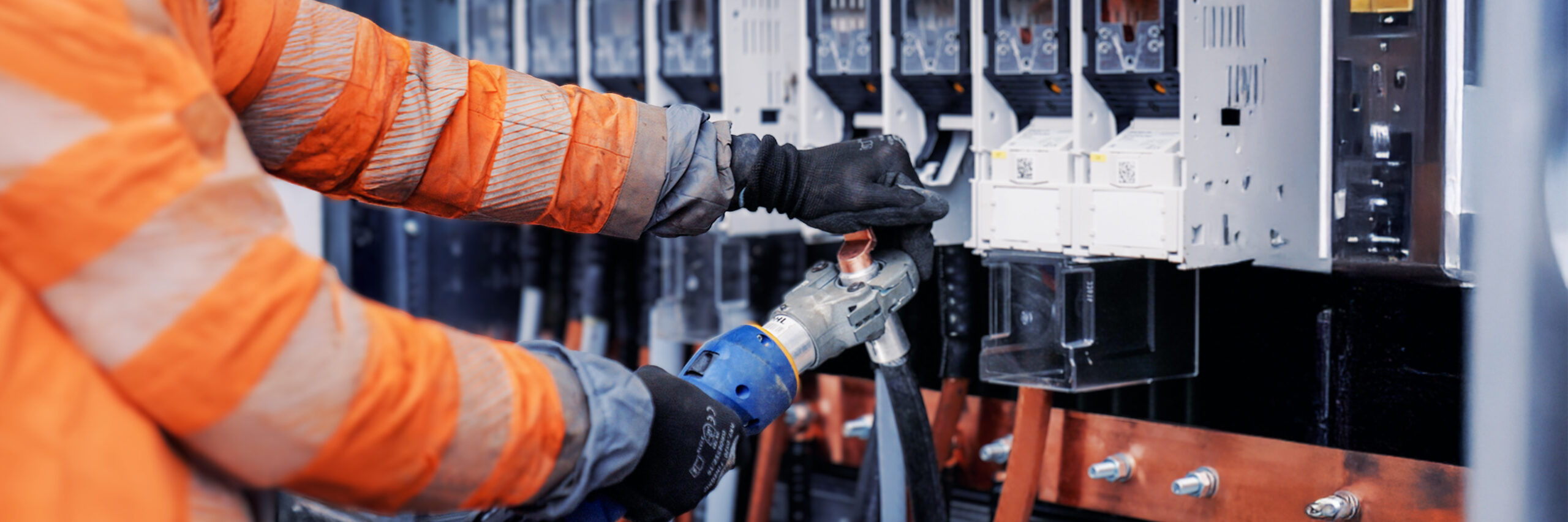 Nahaufnahme von Händen in schwarzen Handschuhen, die mit einem Spezialwerkzeug dicke Kabel an einem Schaltschrank bearbeiten. Die Person trägt eine orangene Pohl-Schutzjacke.